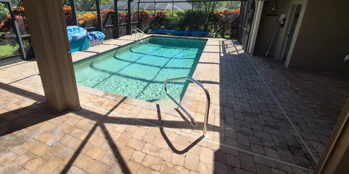 Before: a screened pool in Kings Lake, Naples, surrounded by a tan and brown concrete paver deck with dark staining across the covered lanai