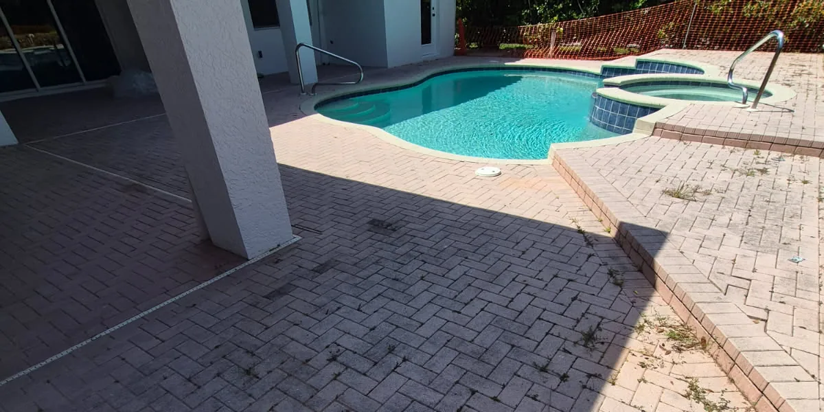 Before: a pool and spa at a home in The Vineyards, Naples, with a weathered herringbone concrete paver deck, weeds growing in the joints and dark staining under the lanai
